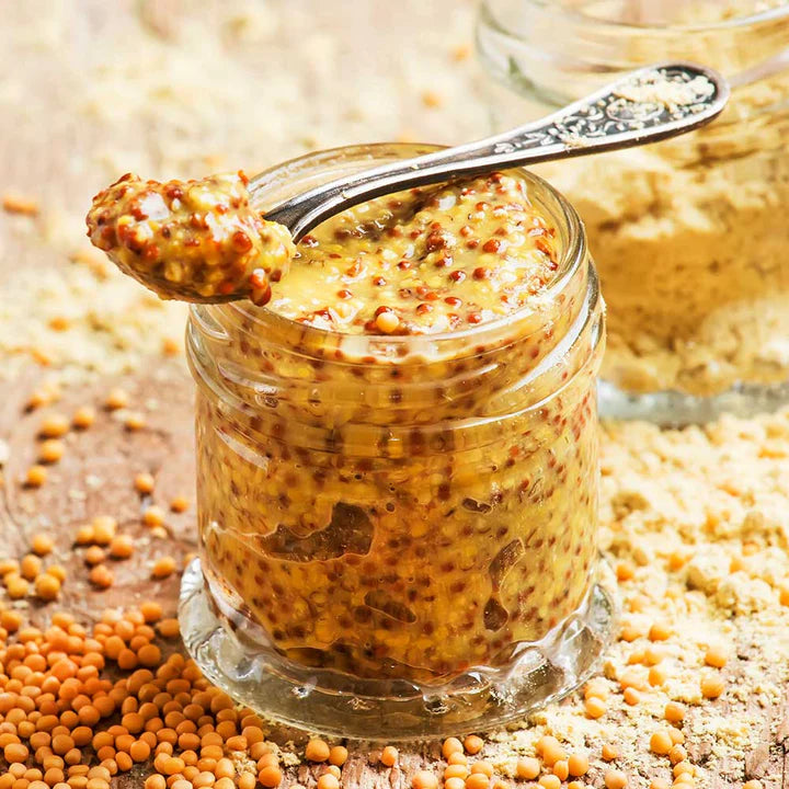 Jar of mustard with a spoonful on top, surrounded by mustard seeds.