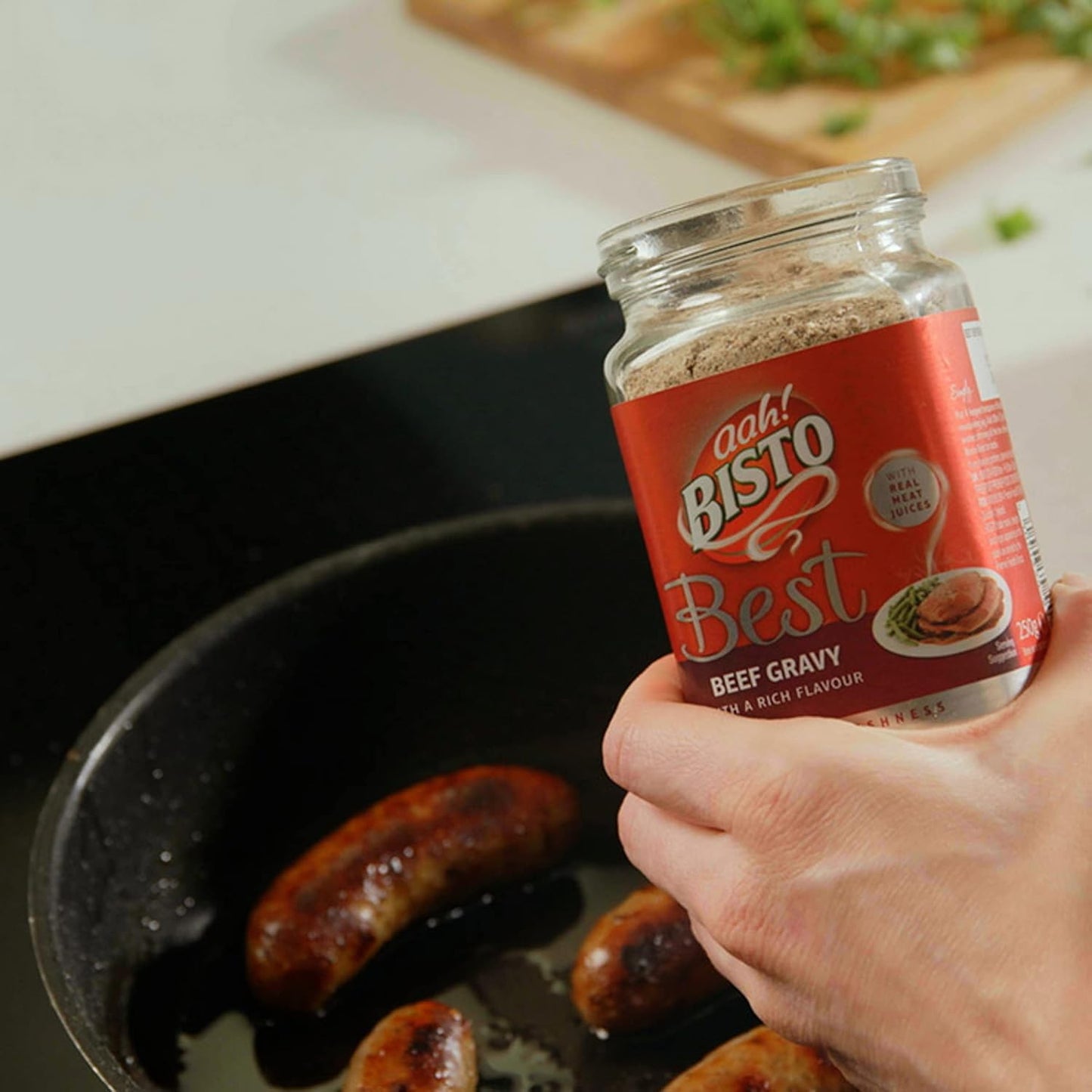 Person holding a jar of Bisto Best Beef Gravy over a pan with sausages.