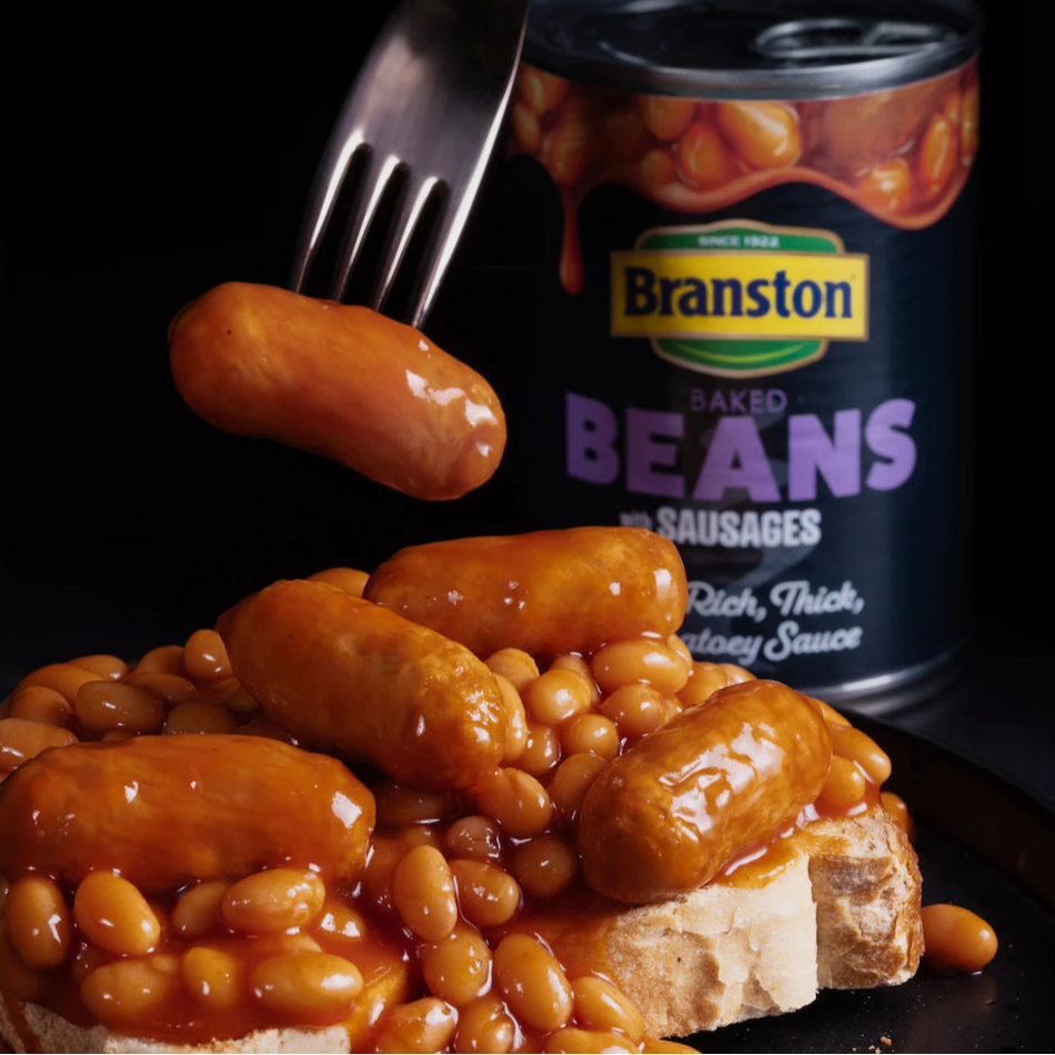 Baked beans with sausages on a bun next to a can of Branston baked beans.