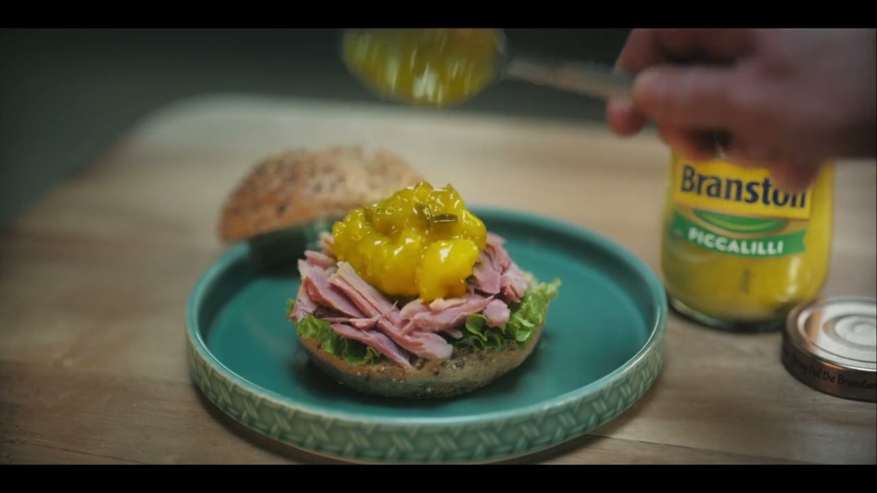 Person adding piccalilli to a sandwich on a wooden board with a jar of Branston Pickle visible.