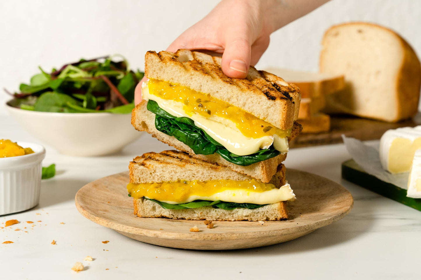 Hand holding a grilled sandwich with egg and spinach on a wooden plate, with bread and salad in the background.