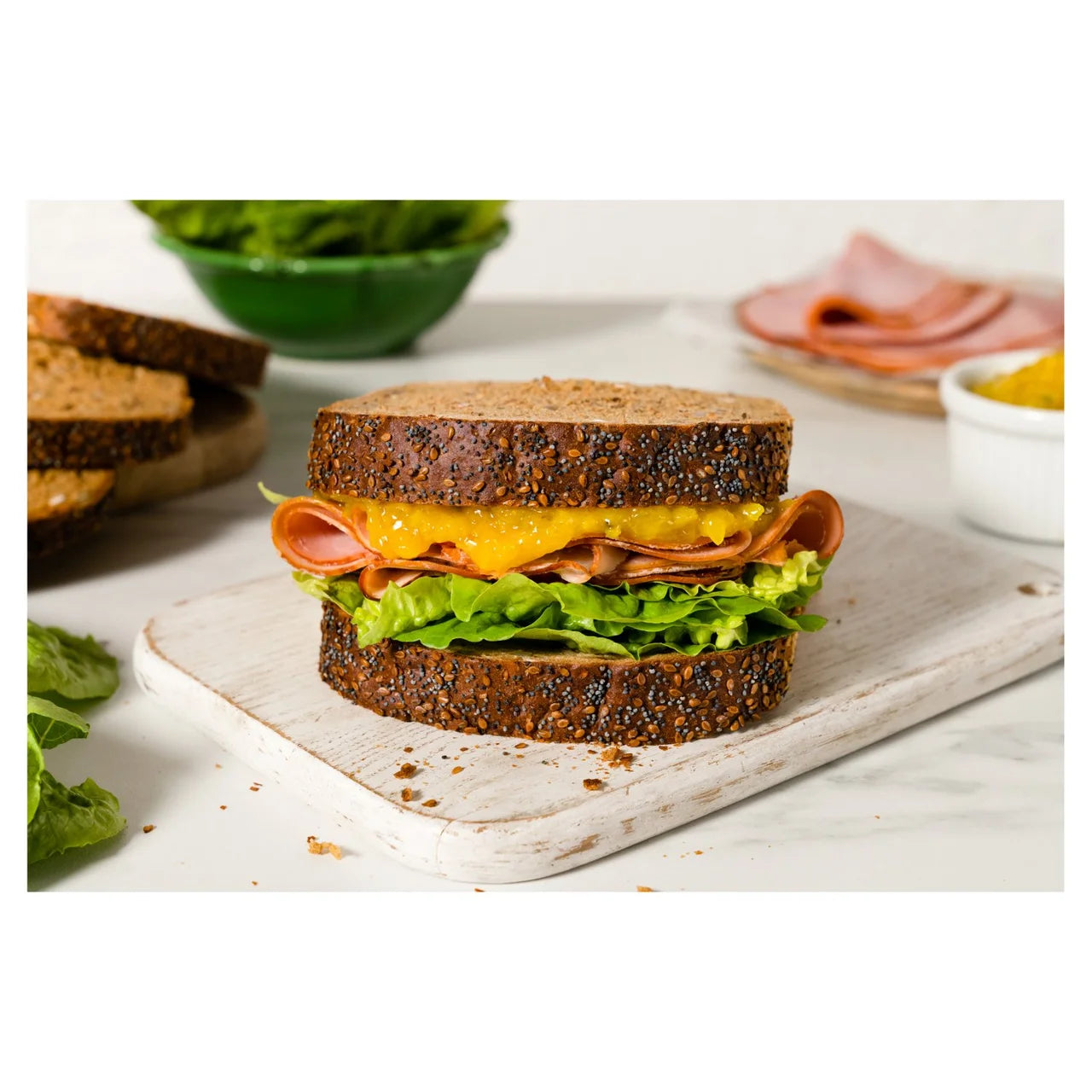 Sandwich with lettuce, tomato, and cheese on a wooden board with ingredients in the background.