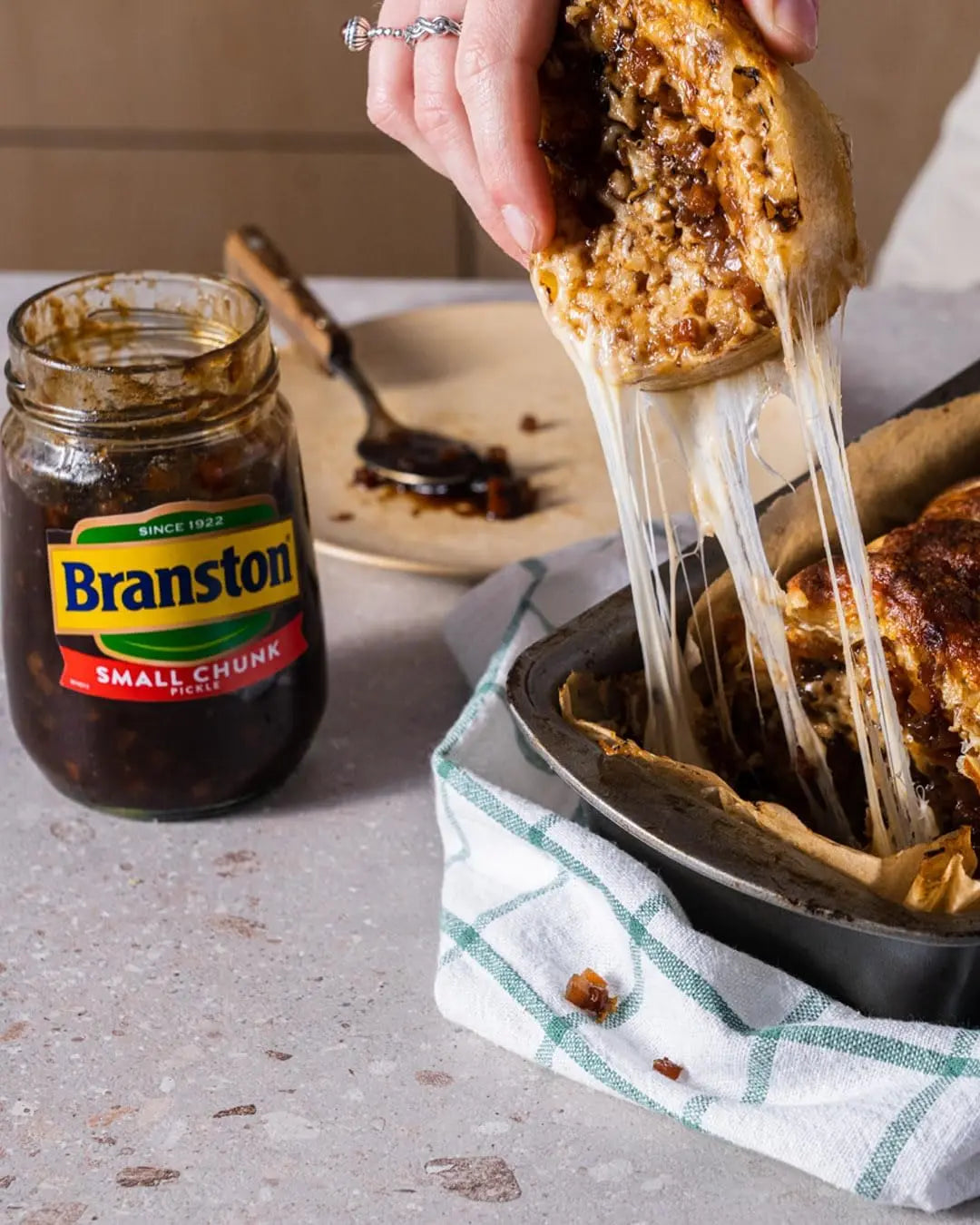 Person holding a piece of food with Branston pickle being pulled, jar of Branston pickle on a table.