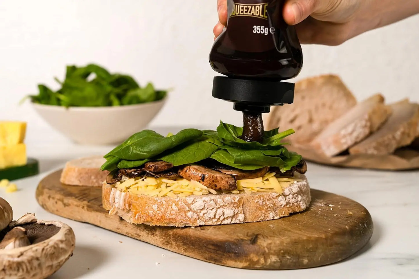 Person adding a bottle of sauce to a sandwich with spinach on a wooden board.