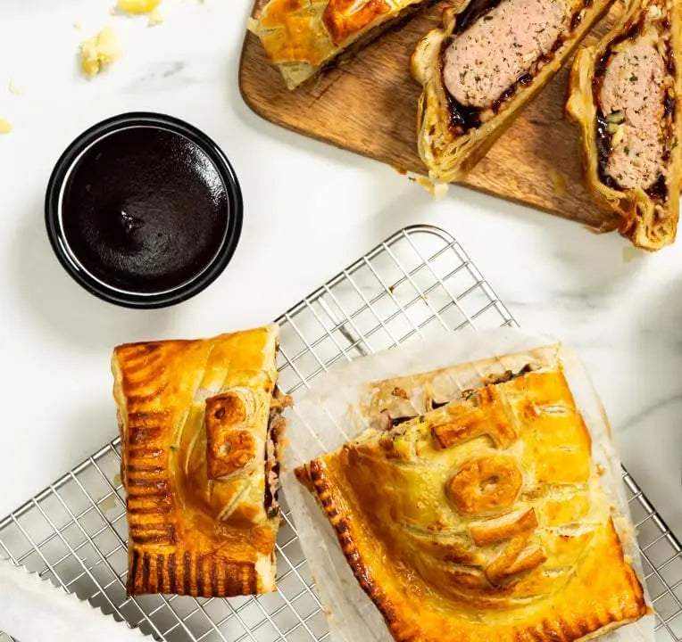 Pork pies on a cooling rack with a small bowl of sauce on a white surface
