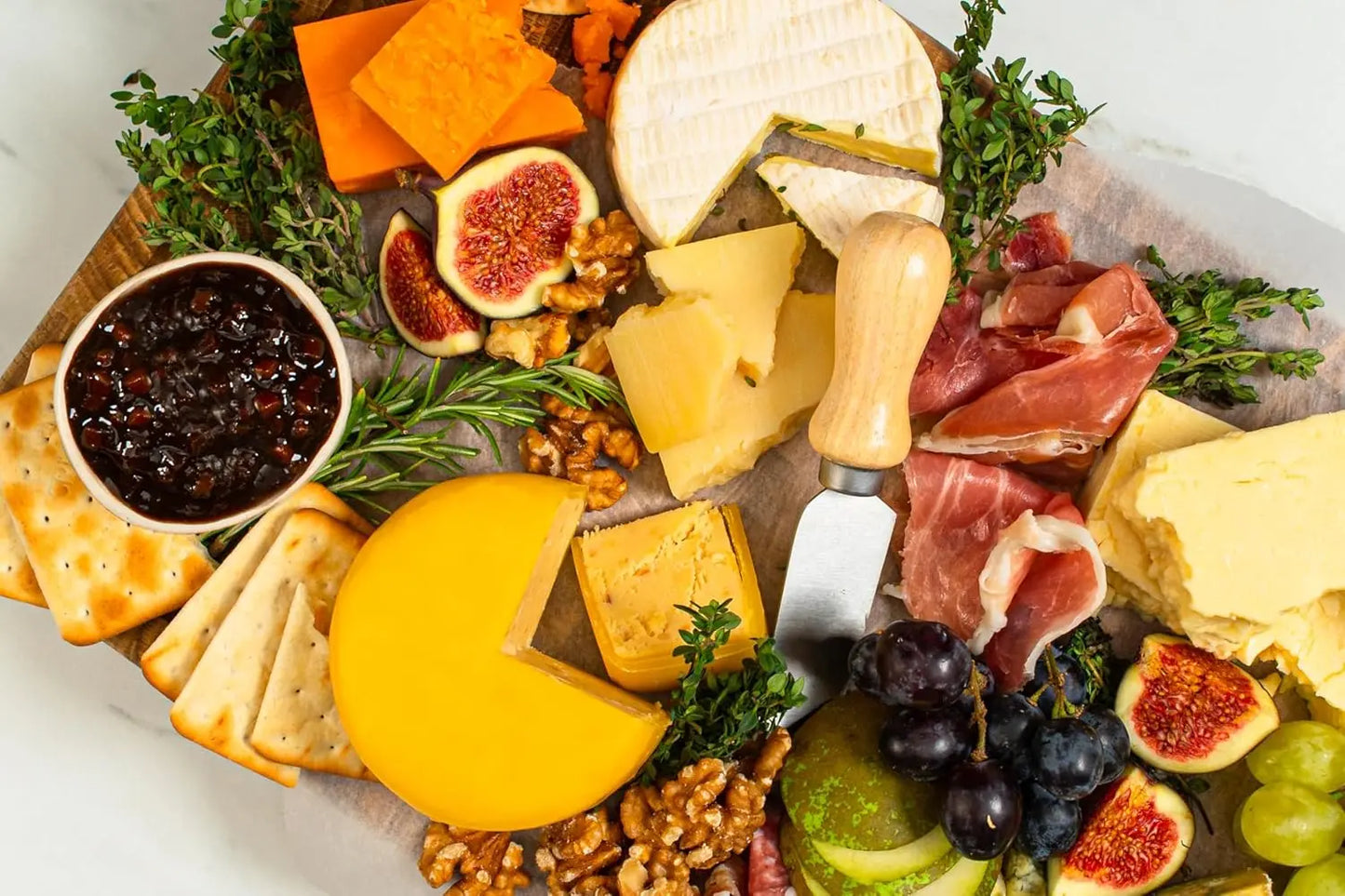 Assorted cheese, fruits, and meats on a wooden board with a knife.