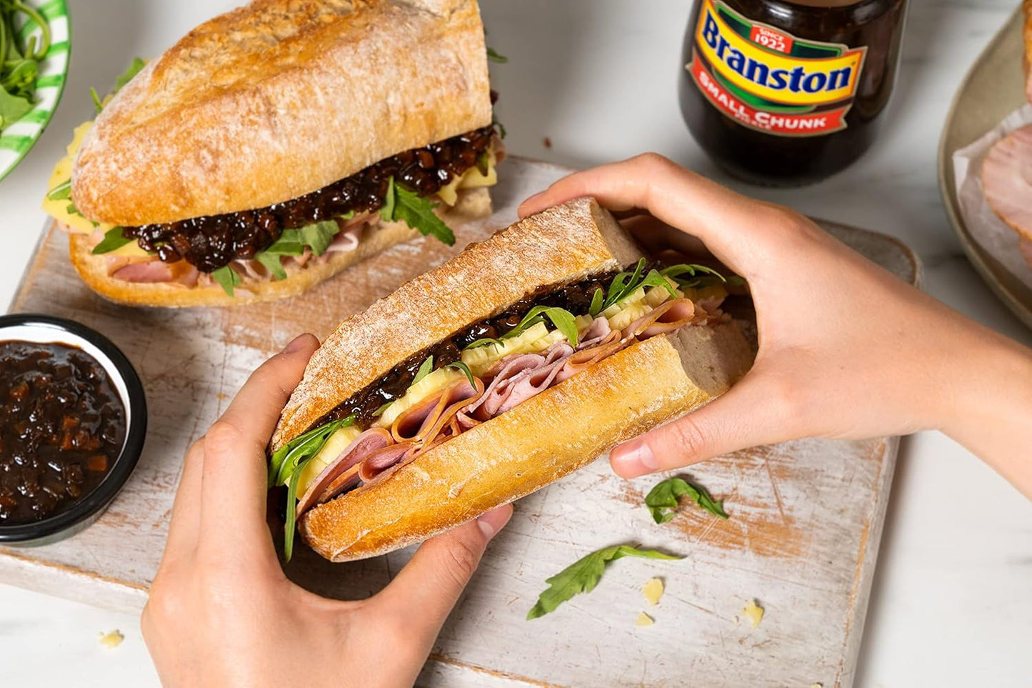 Submarine sandwich being held by hands with Branston pickle in the background.