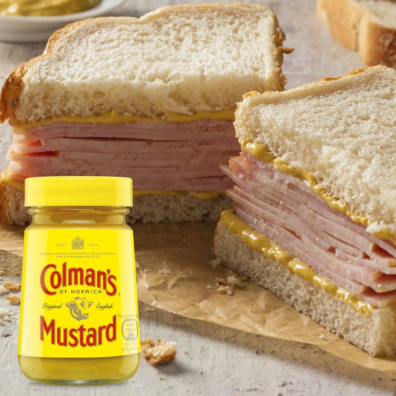 Two sandwiches with ham and mustard, one cut open, on a wooden board with a jar of Colman's Mustard.