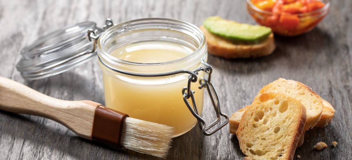 Jar of butter with a wooden brush on a wooden surface with bread slices and a bowl of vegetables.