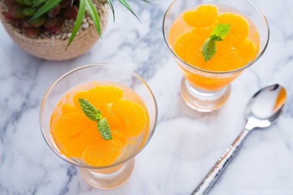 Two glass dishes with orange dessert on a marble surface with a spoon and plant.