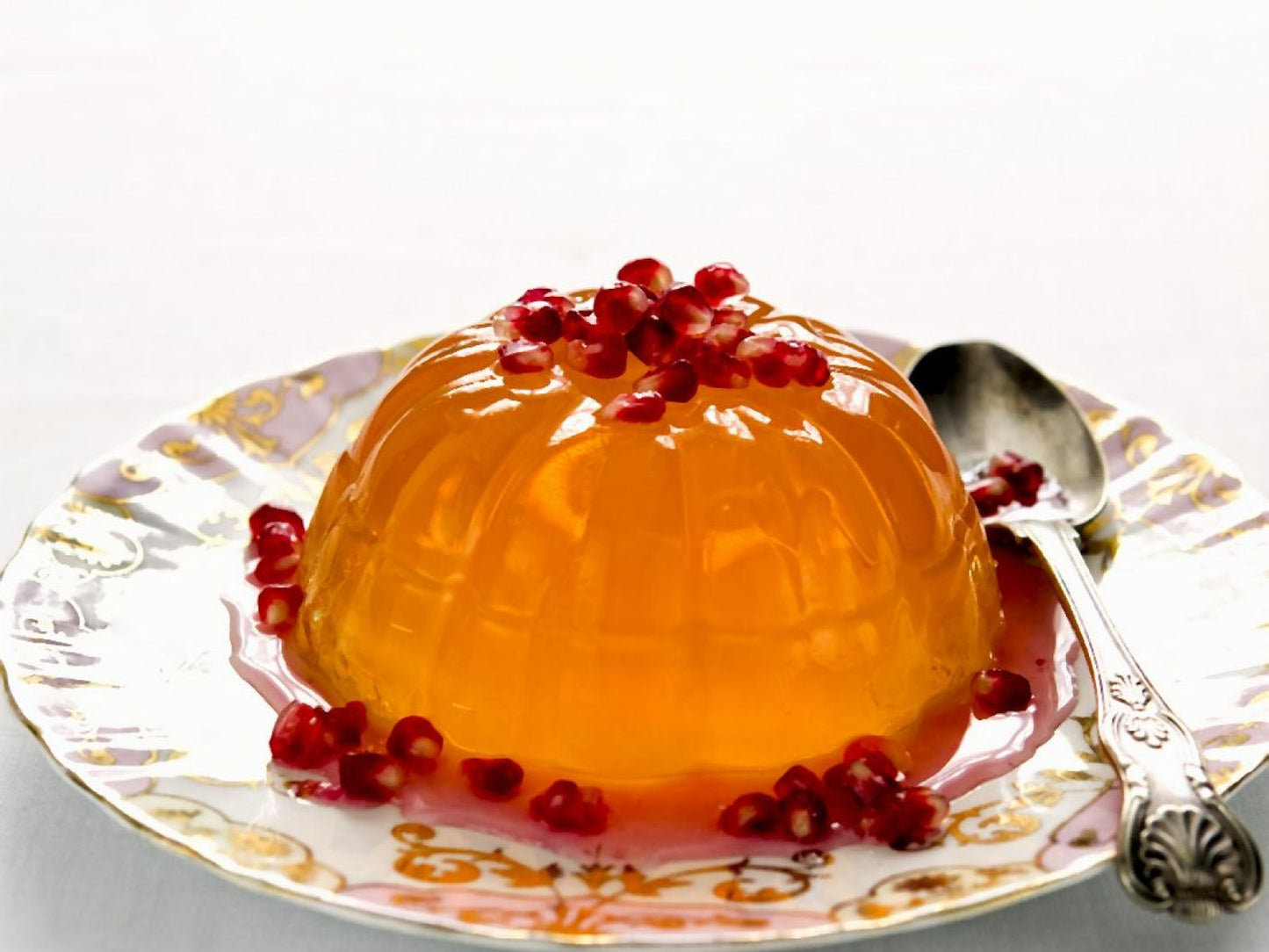 Orange jelly dessert with pomegranate seeds on a decorative plate with a spoon.