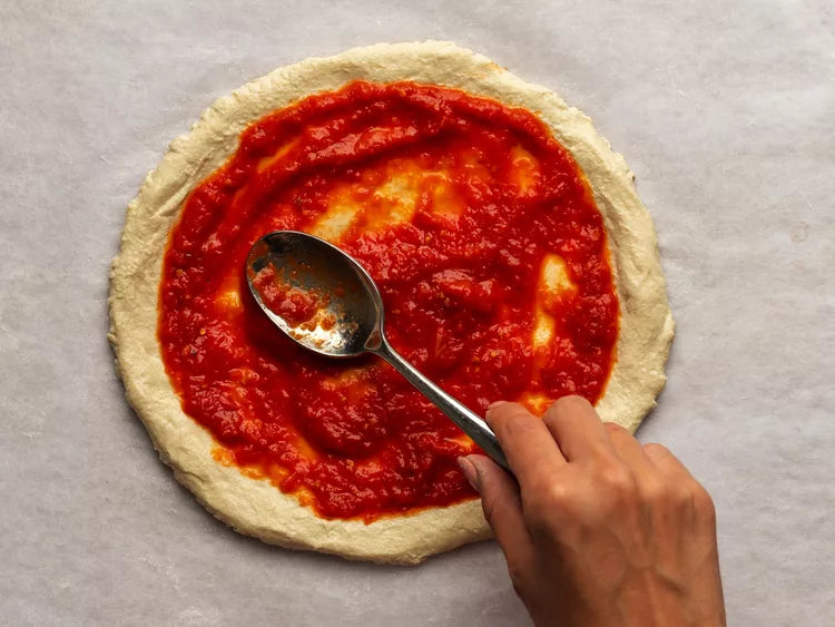 Person spreading tomato sauce on a pizza dough with a spoon on a gray surface.