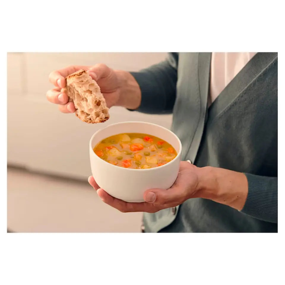 Person holding a white bowl of vegetable soup with visible carrots and celery, dipping a piece of rustic bread into the soup.