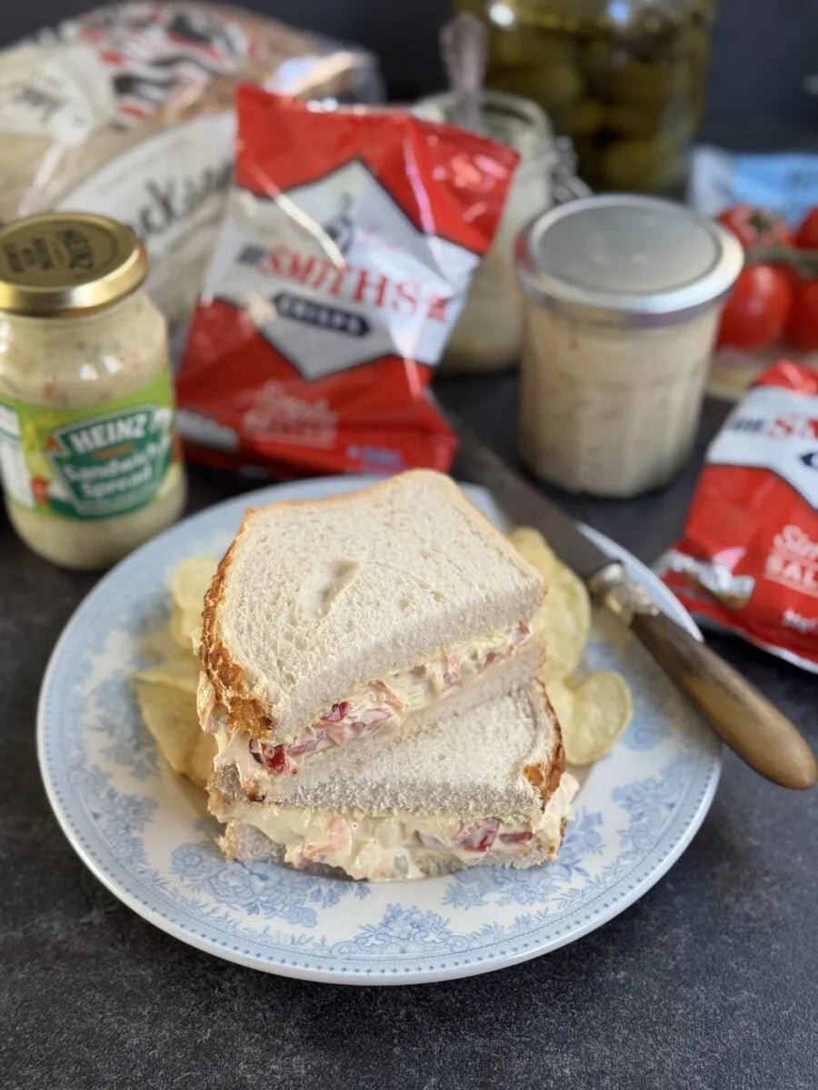 Sandwich on a plate with chips, surrounded by condiment jars and a Smith's packet on a dark surface.