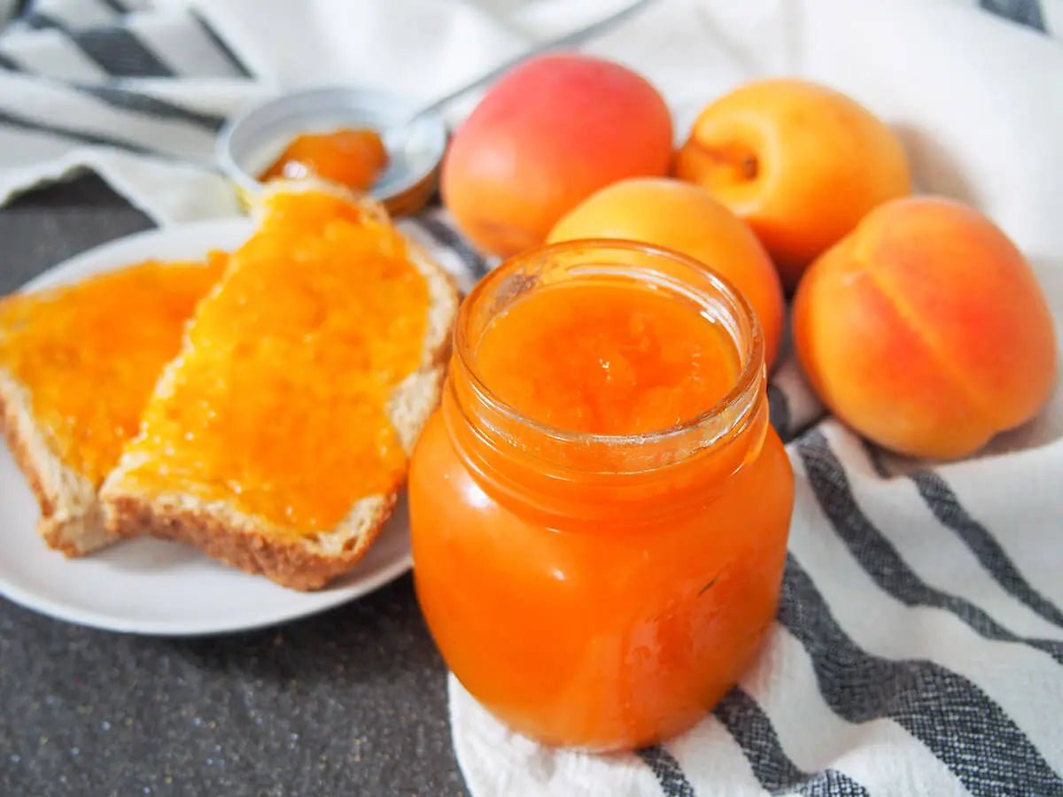 Jar of apricot jam with sliced bread and fresh apricots on a striped cloth.