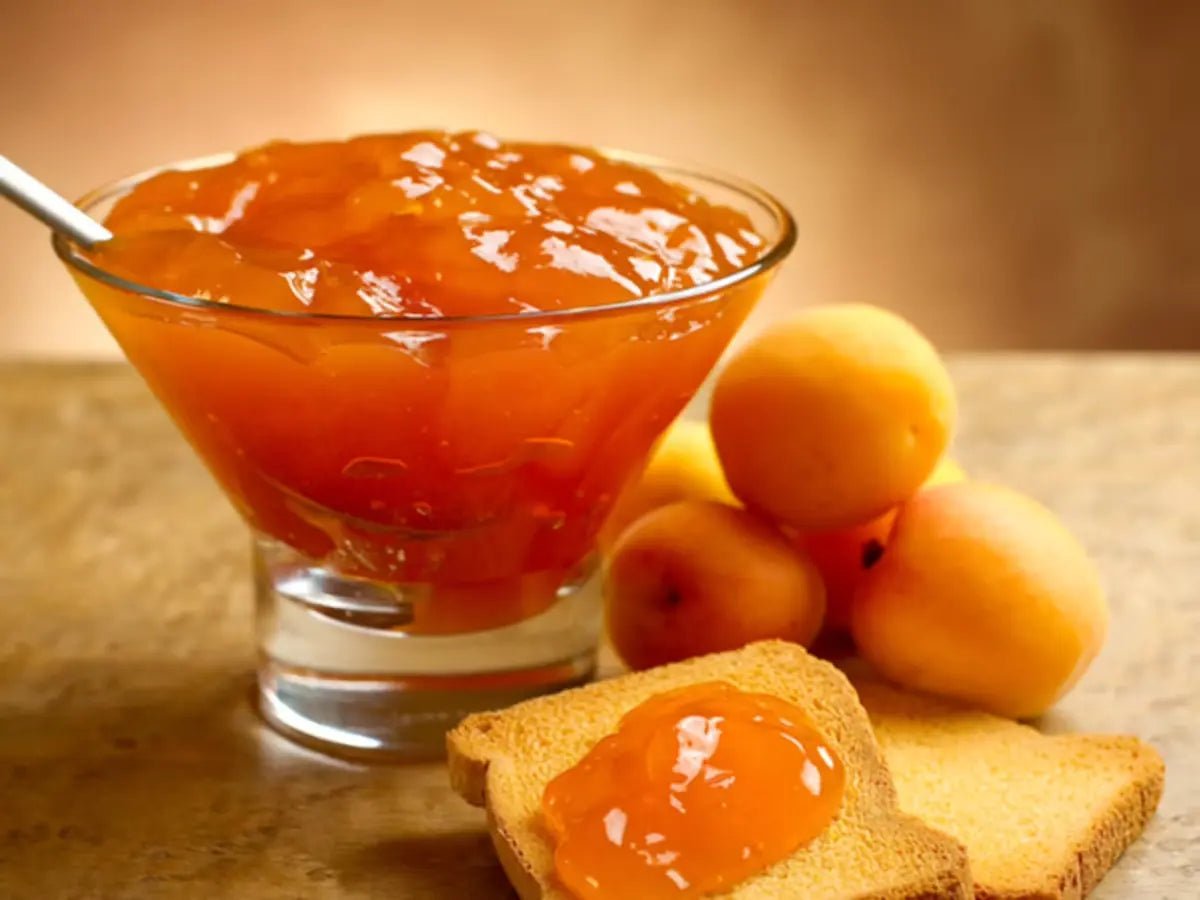 Glass bowl of apricot jam with a spoon, surrounded by apricots and toast on a wooden surface.