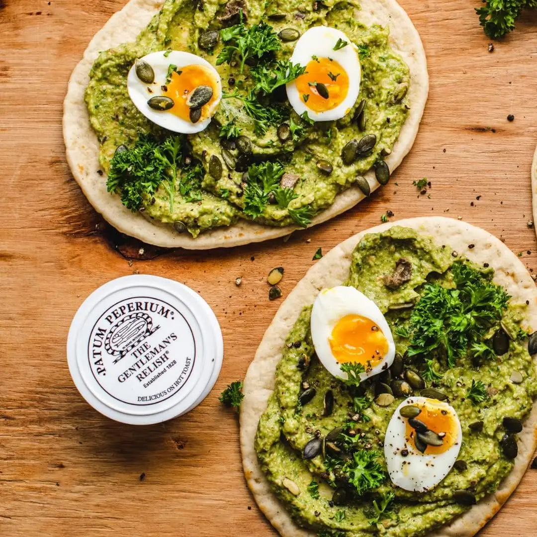 Two flatbreads with green spread, boiled eggs, and herbs on a wooden surface with a jar of Saturey Peperium relish.