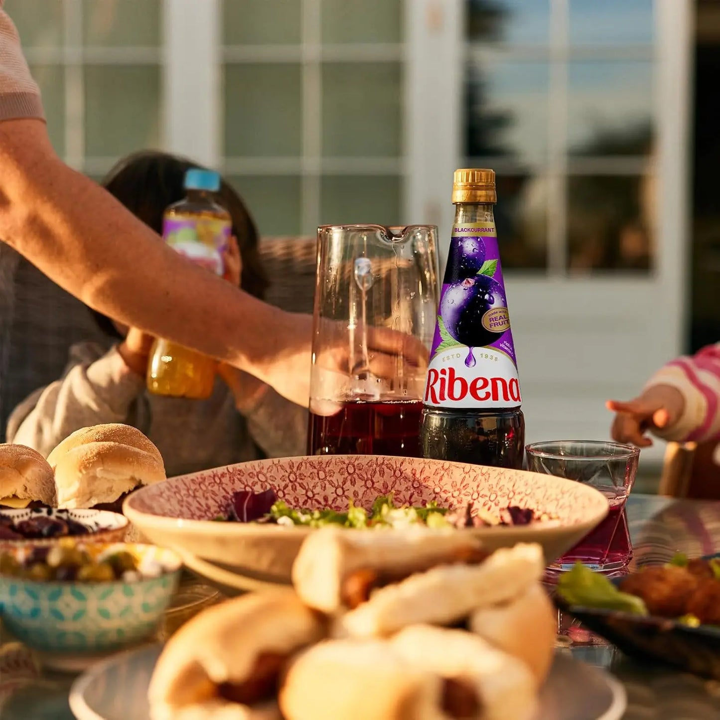 Table with food and drinks, including a bottle of Ribena, with people in the background.