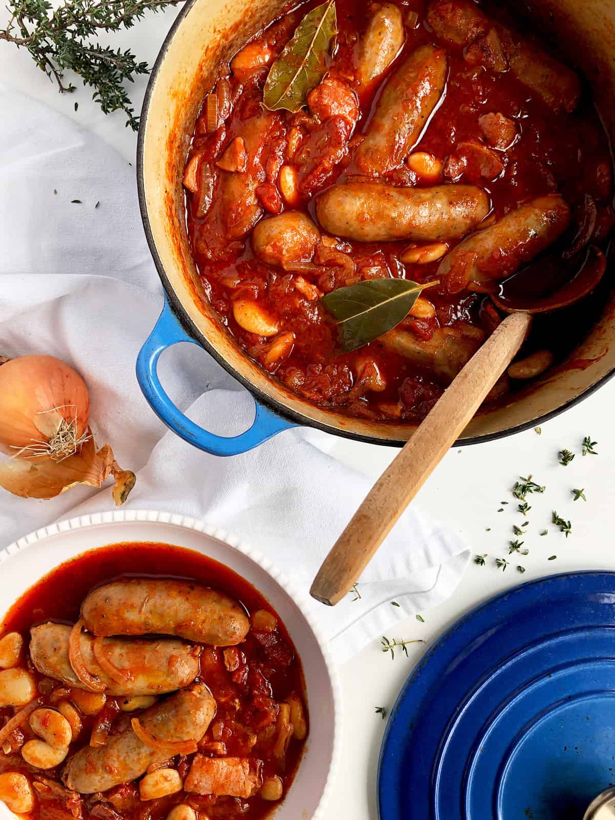 Pot of sausage and bean stew with a wooden spoon, surrounded by ingredients on a white surface.