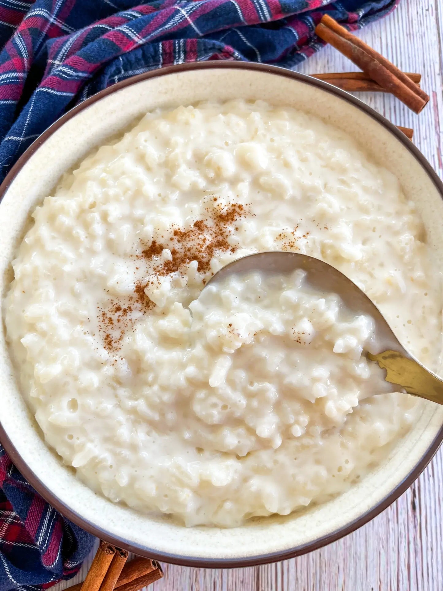 Bowl of rice pudding with a spoon, cinnamon, and plaid cloth on a wooden surface