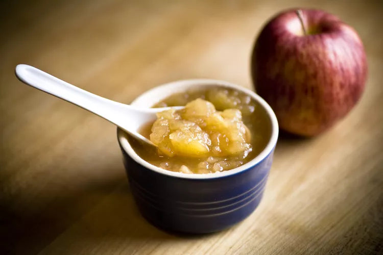Bowl of applesauce with a spoon and an apple on a wooden surface
