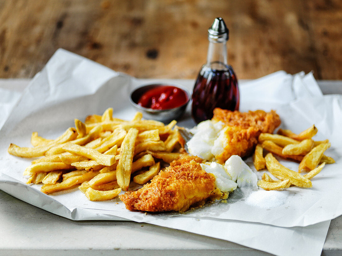 Fish and chips served on a white plate with ketchup and vinegar bottles in the background.