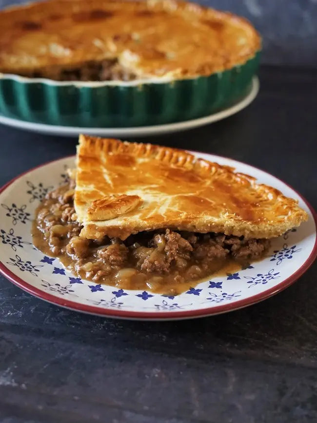 Slice of meat pie on a decorative plate with the rest of the pie in the background.