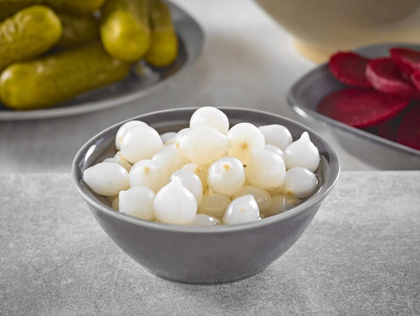 Bowl of white pickled pearl onions on a gray surface with pickles in the background