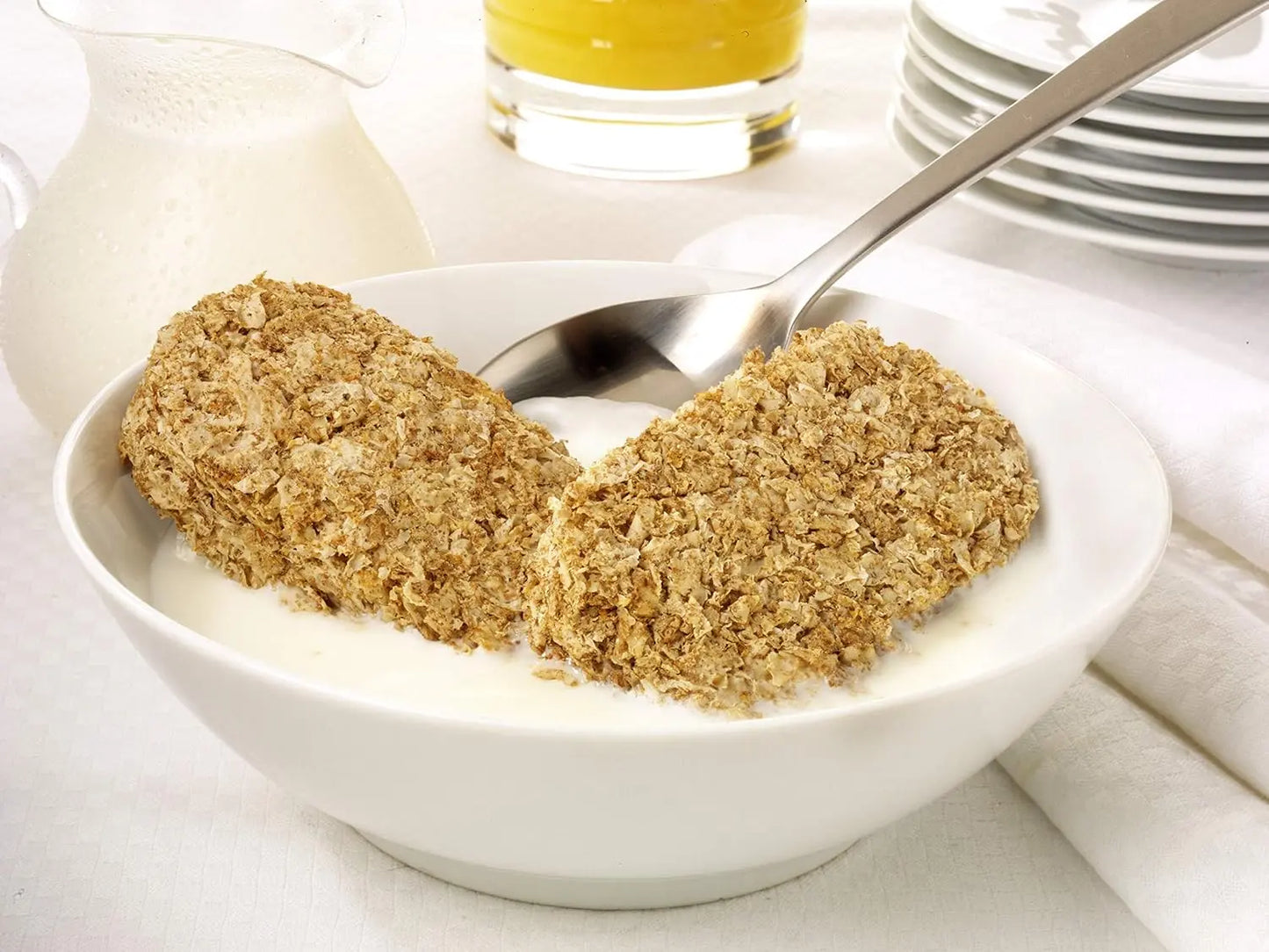Two oatmeal cookies in a bowl with a spoon, surrounded by a glass of orange juice and a pitcher on a white surface.