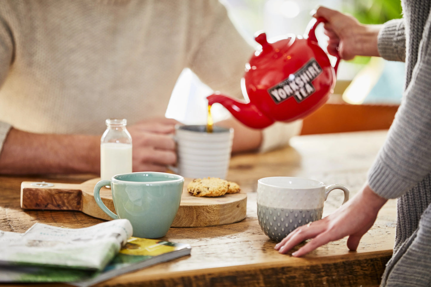 Person pouring Yorkshire Tea into a mug on a wooden table with another person holding a cookie.