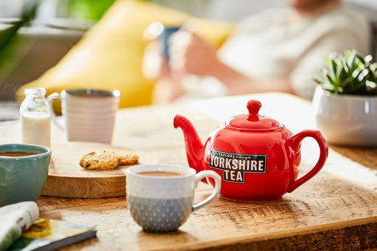 A red Yorkshire Tea teapot with a cup of tea, milk jug, and biscuits on a wooden table, creating a cosy British tea scene.