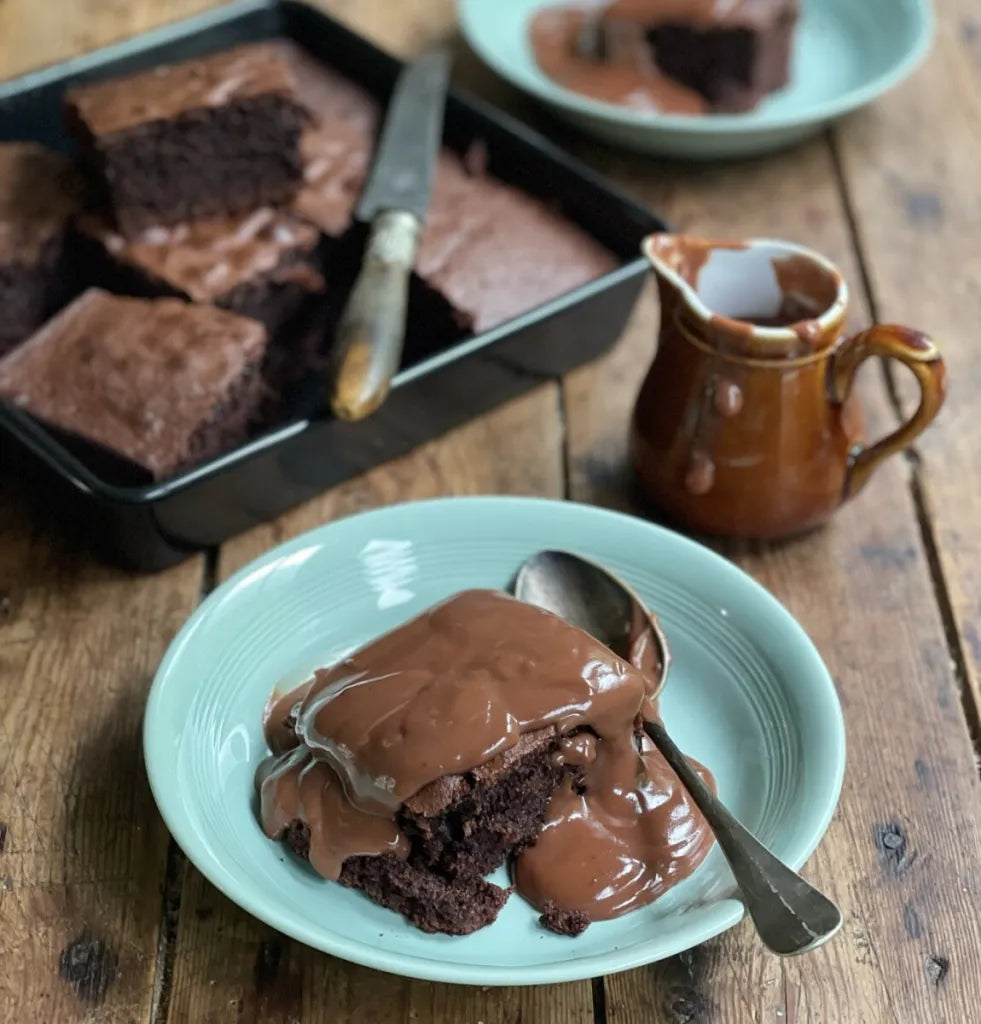 Chocolate brownie with a scoop of ice cream on a teal plate, served with a small pitcher of chocolate sauce on a wooden table.