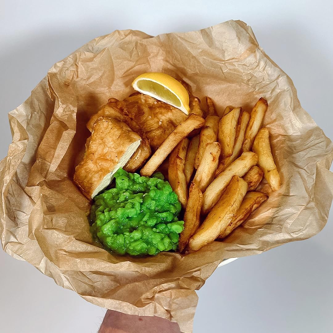 Bowl of fish, chips, and mushy peas on brown paper with a lemon wedge.