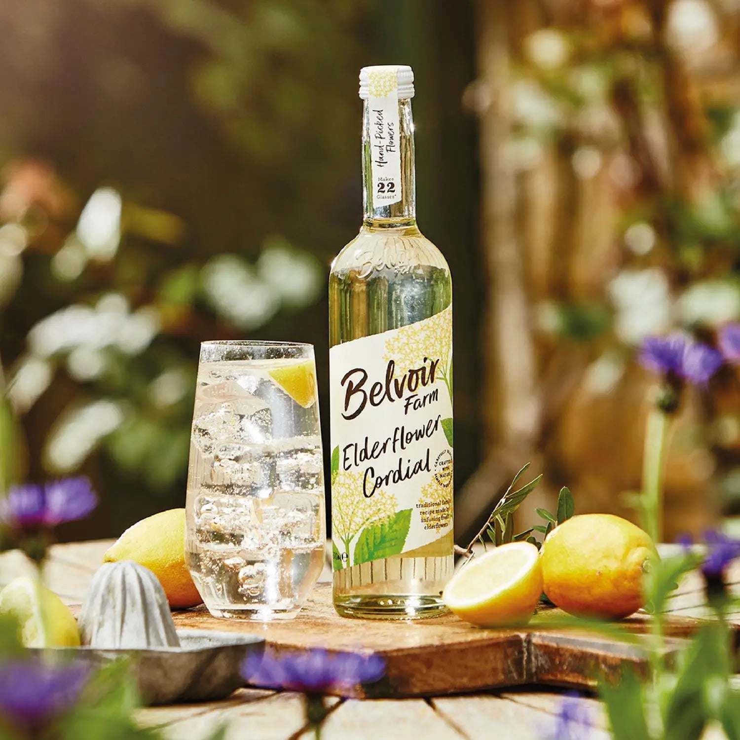 Bottle of Belvoir Farm Elderflower Cordial with a glass of lemonade on a wooden surface outdoors.