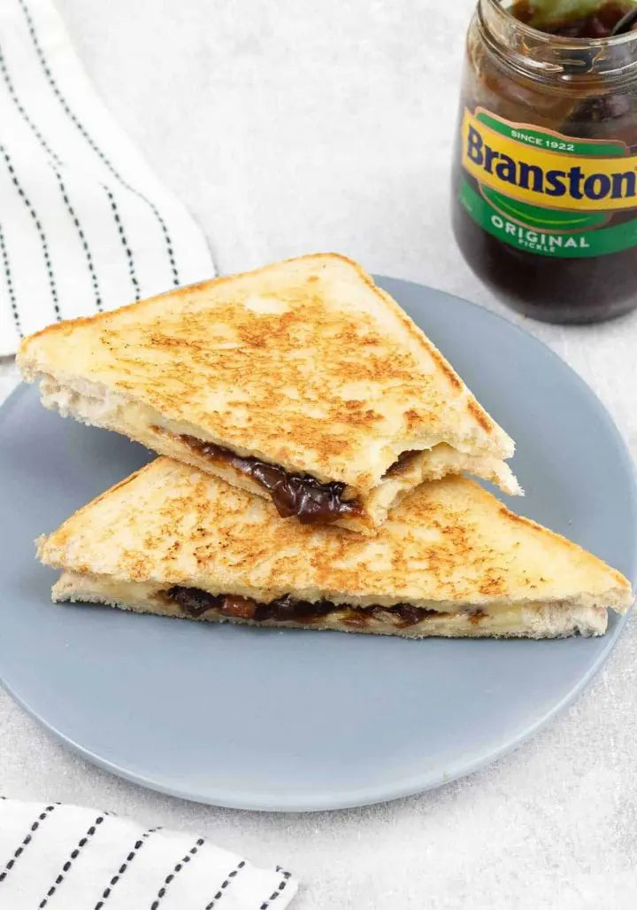 Grilled sandwich with Branston pickle on a blue plate, jar of Branston pickle in the background.