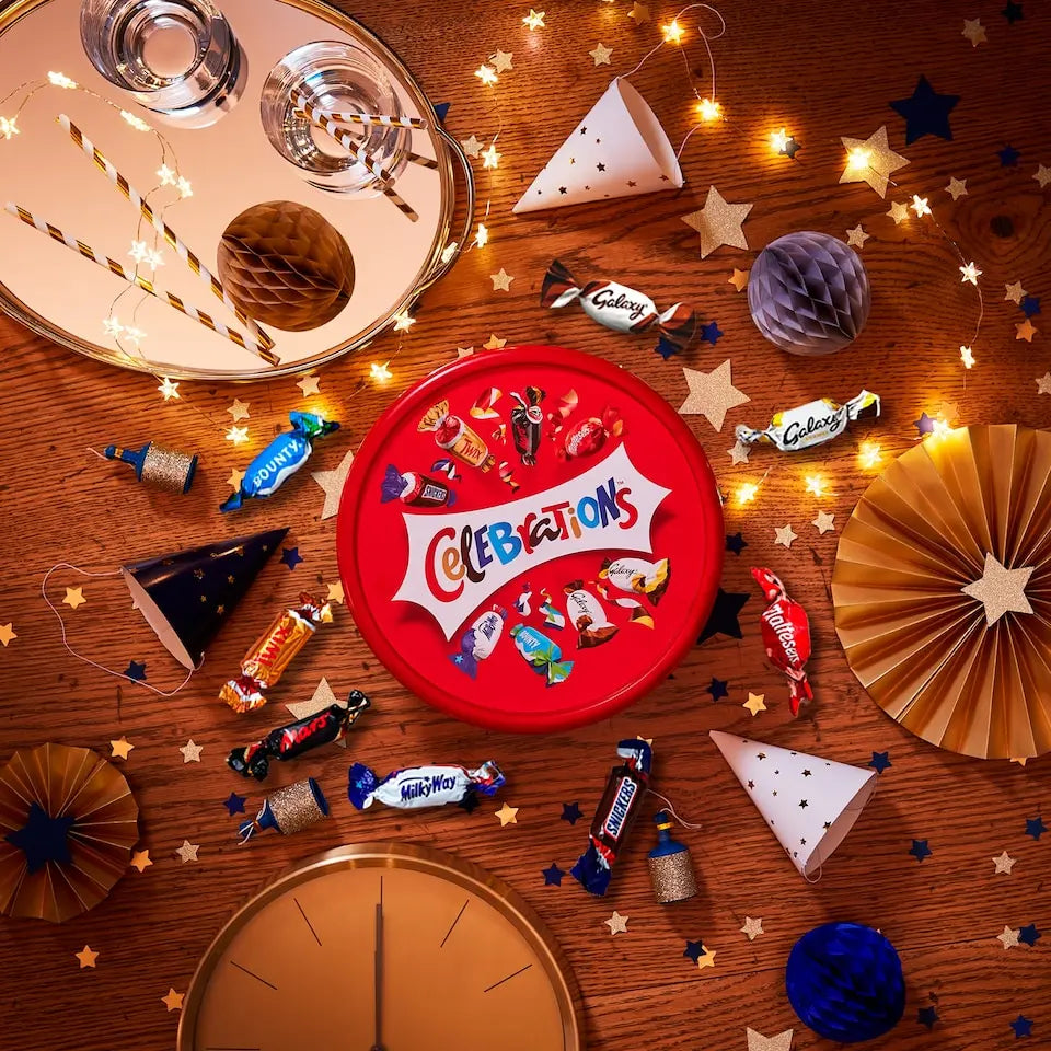 Red 'Celebrations' candy tin surrounded by various candies on a wooden surface with decorative lights and paper fans.