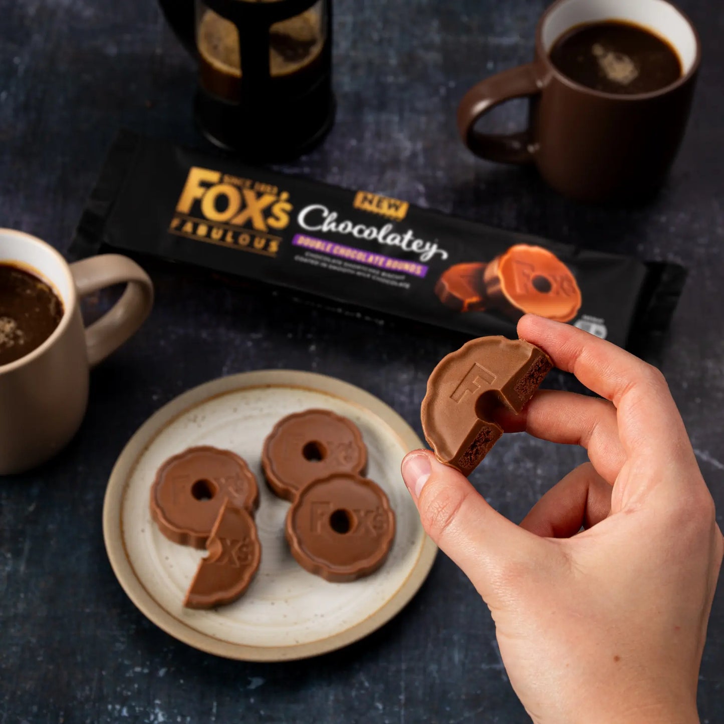 Hand holding a chocolate biscuit with 'Fox's' packaging and mugs of coffee in the background.