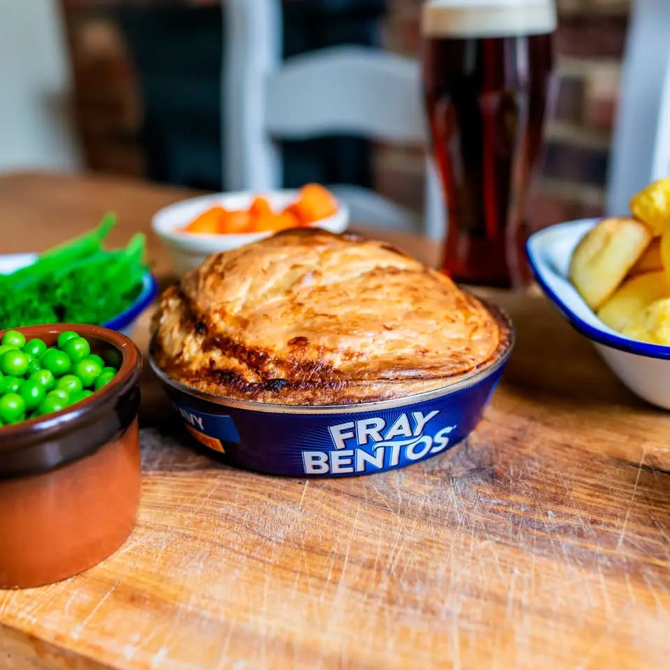 Fray Bentos bento box with a pie on a wooden table with side dishes and a beer.