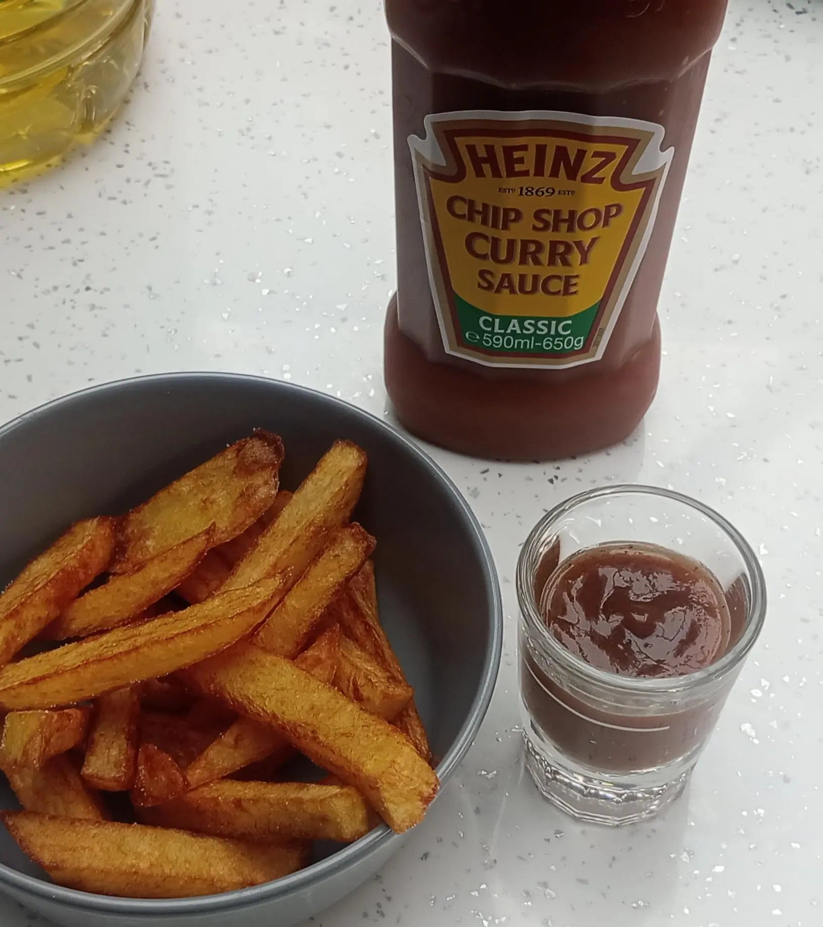 Bowl of fries with a bottle of Heinz Chip Shop Curry Sauce and a small glass of sauce on a white surface.