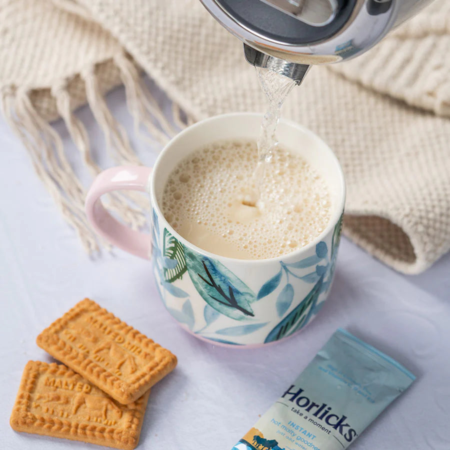 Horlicks instant breakfast drink being poured into a mug with cookies and a textured blanket in the background.