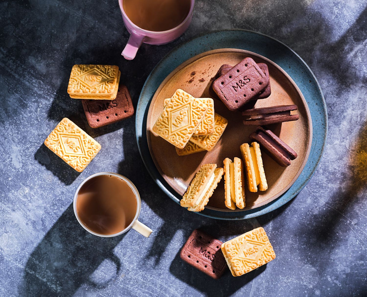Assorted M&S biscuits on a plate with two cups of coffee on a dark surface