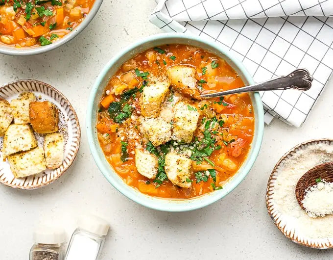 A top-down view of a light blue bowl filled with a chunky vegetable soup, garnished with fresh parsley and grated cheese, with a spoon inside. The bowl is set on a light gray surface alongside a small plate of croutons, a folded white checkered napkin, and other bowls of food.
