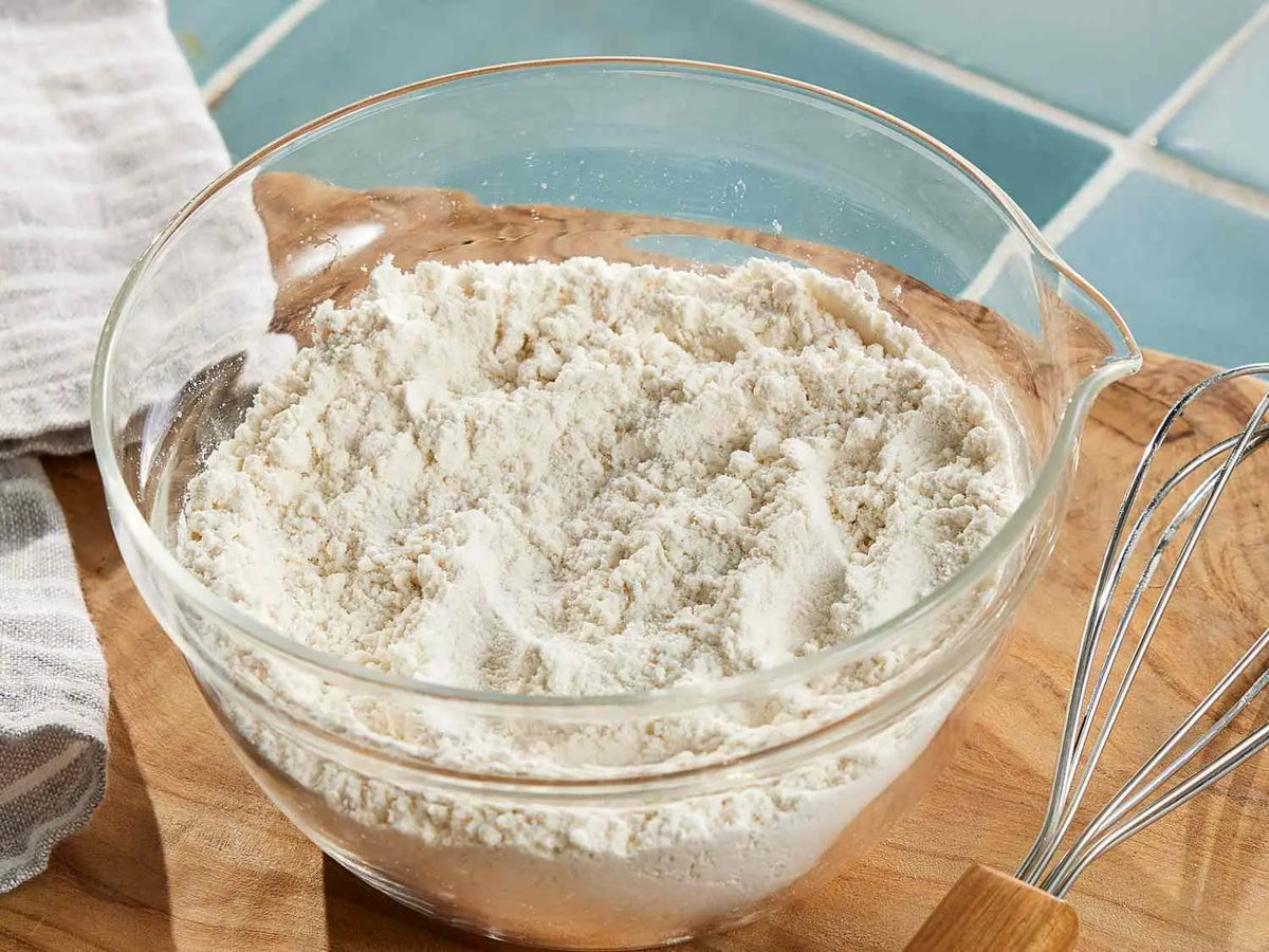 A clear glass bowl filled with white flour, set on a wooden cutting board with a metal whisk nearby and a blue tiled background.