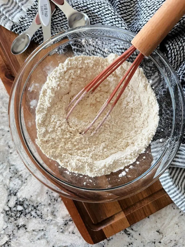 A clear glass mixing bowl filled with flour, with a wooden-handled whisk resting inside. The bowl sits on a wooden cutting board over a speckled stone countertop, with metal measuring spoons and a striped kitchen towel nearby.
