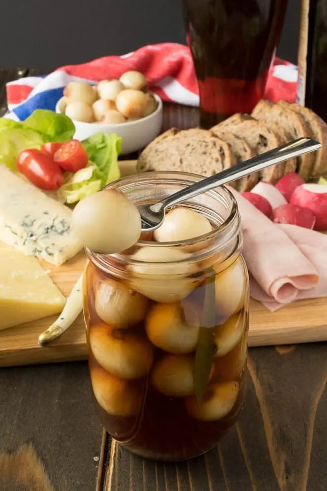 A glass mason jar filled with pickled onions in a dark sauce, set on a rustic wooden table with a spread of sliced bread, cheese, radishes, and a side salad in the background.