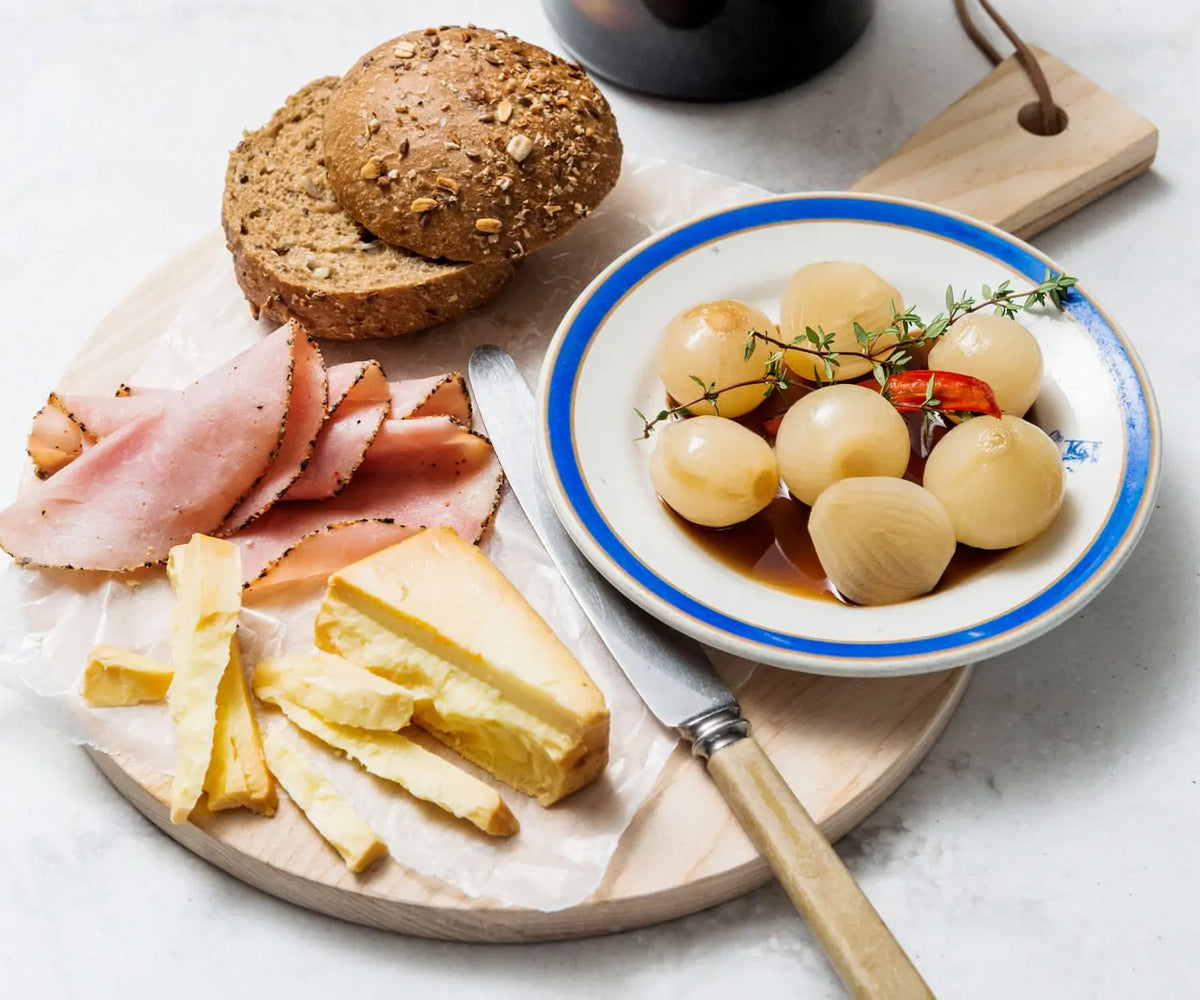 A styled food spread on a round wooden board with sliced bread, ham, cheese, and a bowl of marinated onion or garlic in sauce, photographed on a light stone surface.