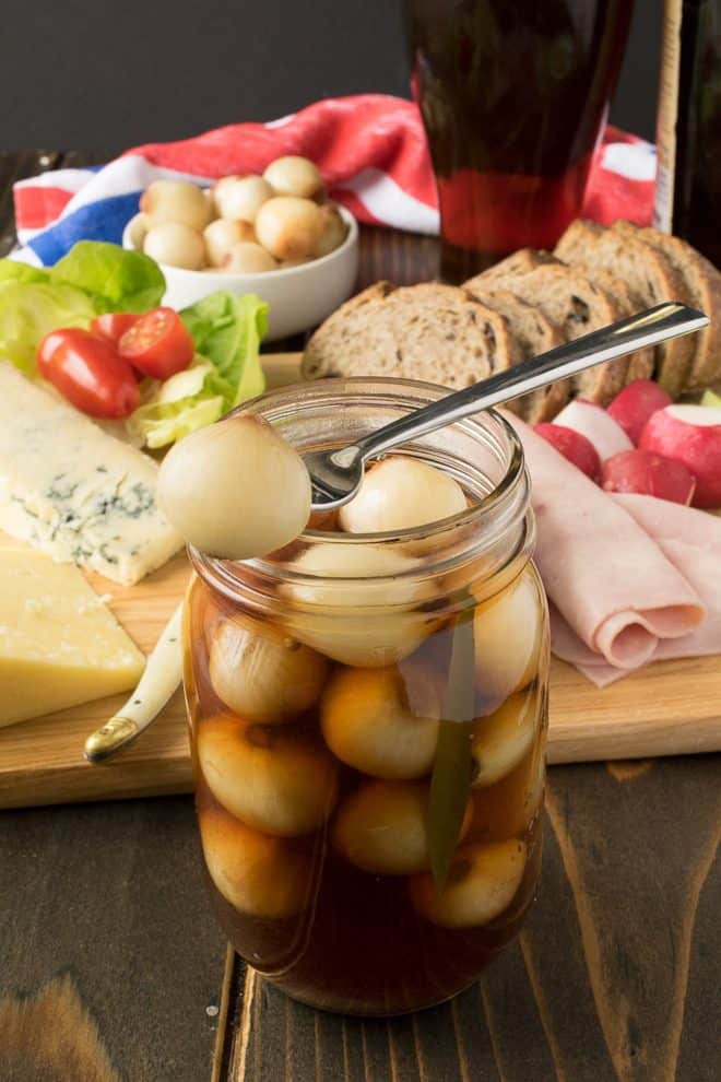 Jar of pickled eggs with a spoon, surrounded by bread, cheese, and vegetables on a wooden table.