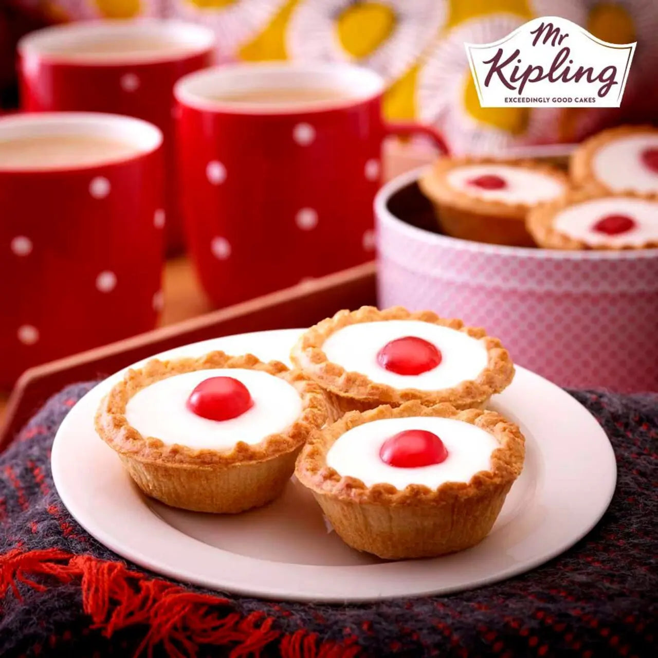 Three small tarts with red centers on a plate with red polka dot mugs and a bowl of more tarts in the background, featuring the Mr Kipling logo.