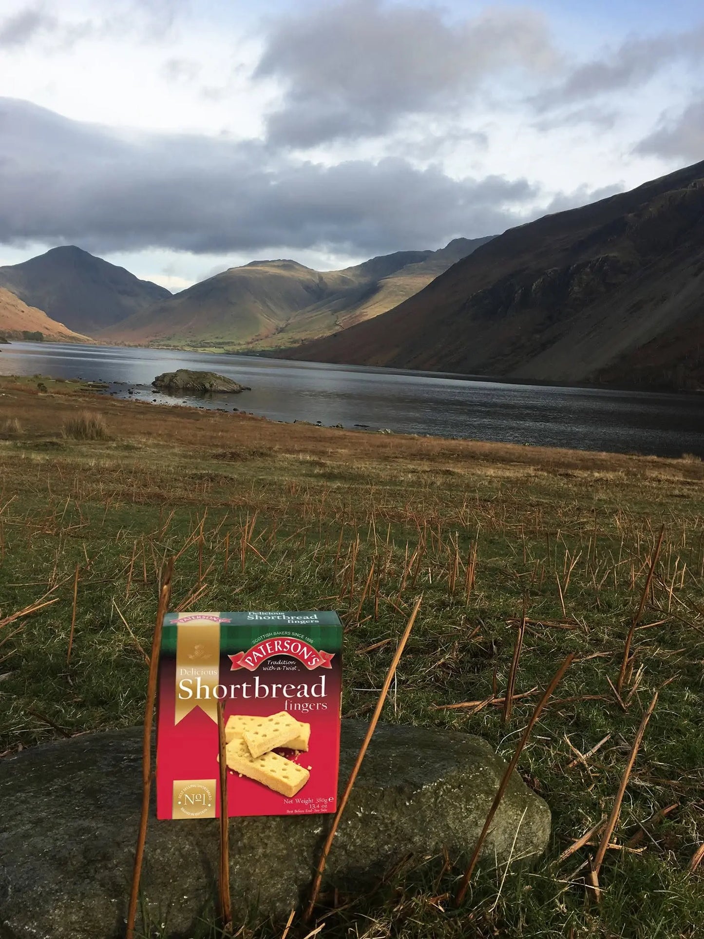 A package of Maitland's Shortbread Biscuits placed on a rock in a grassy field, with a scenic mountain lake landscape in the background under a cloudy sky.
