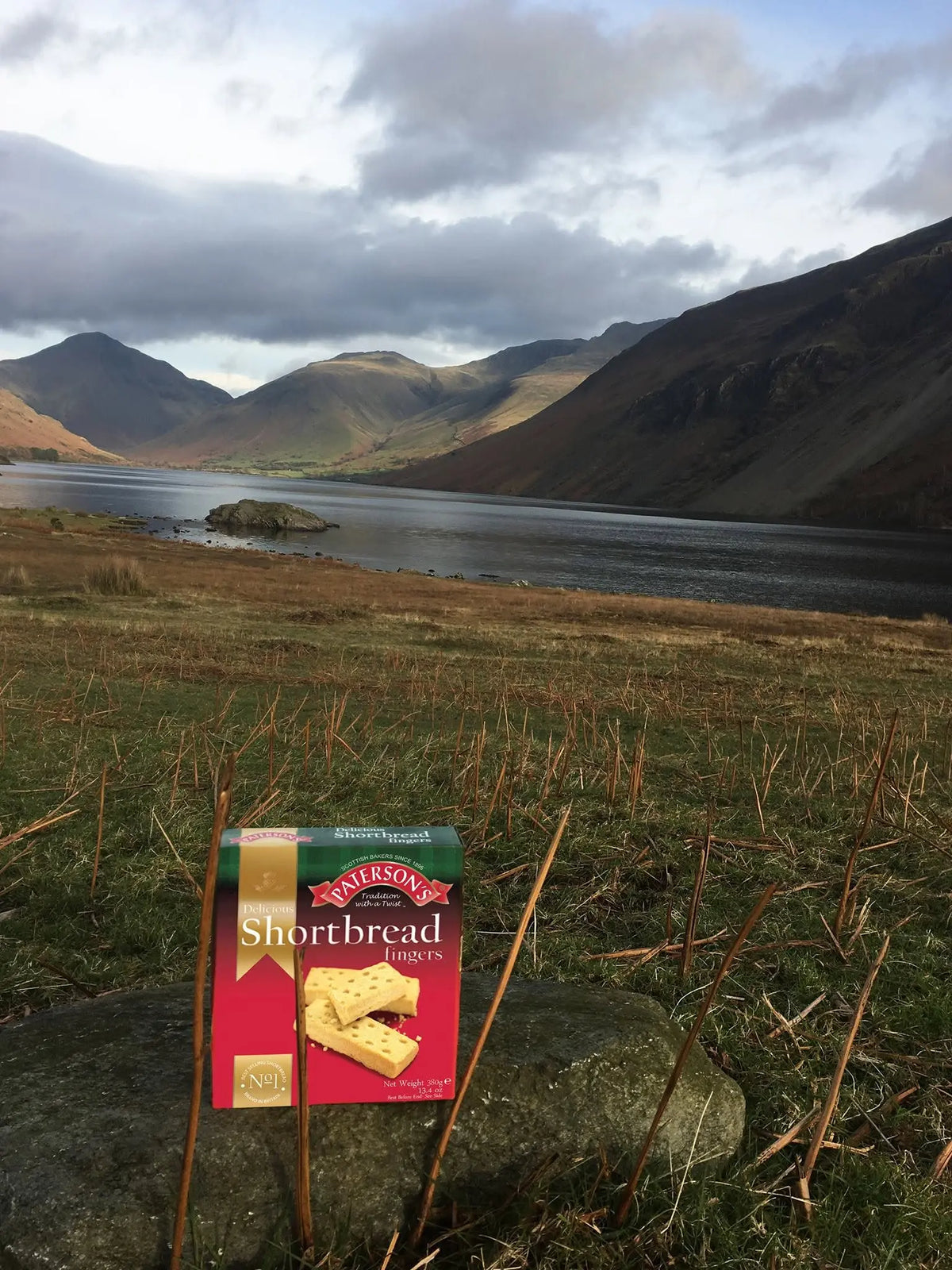 A package of Maitland's Shortbread Biscuits placed on a rock in a grassy field, with a scenic mountain lake landscape in the background under a cloudy sky.